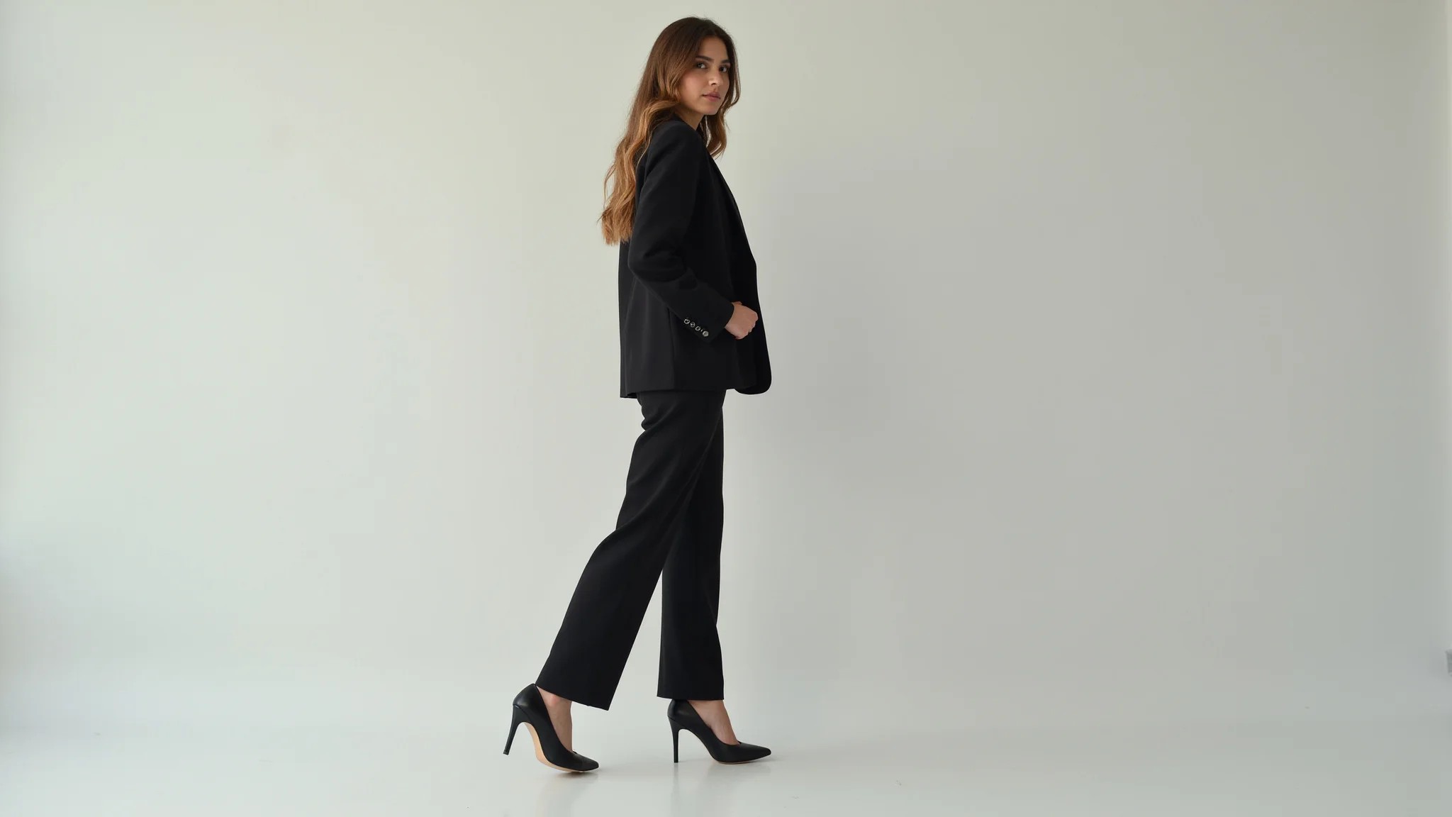 Mulher brasileira vestindo roupa monocromática escura, salto alto, posando em estúdio minimalista.