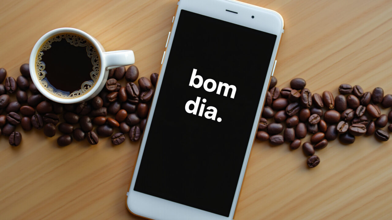 A cup of coffee next to a phone displaying a 'bom dia' message with a coffee bean background. Café com frase de bom dia com café.